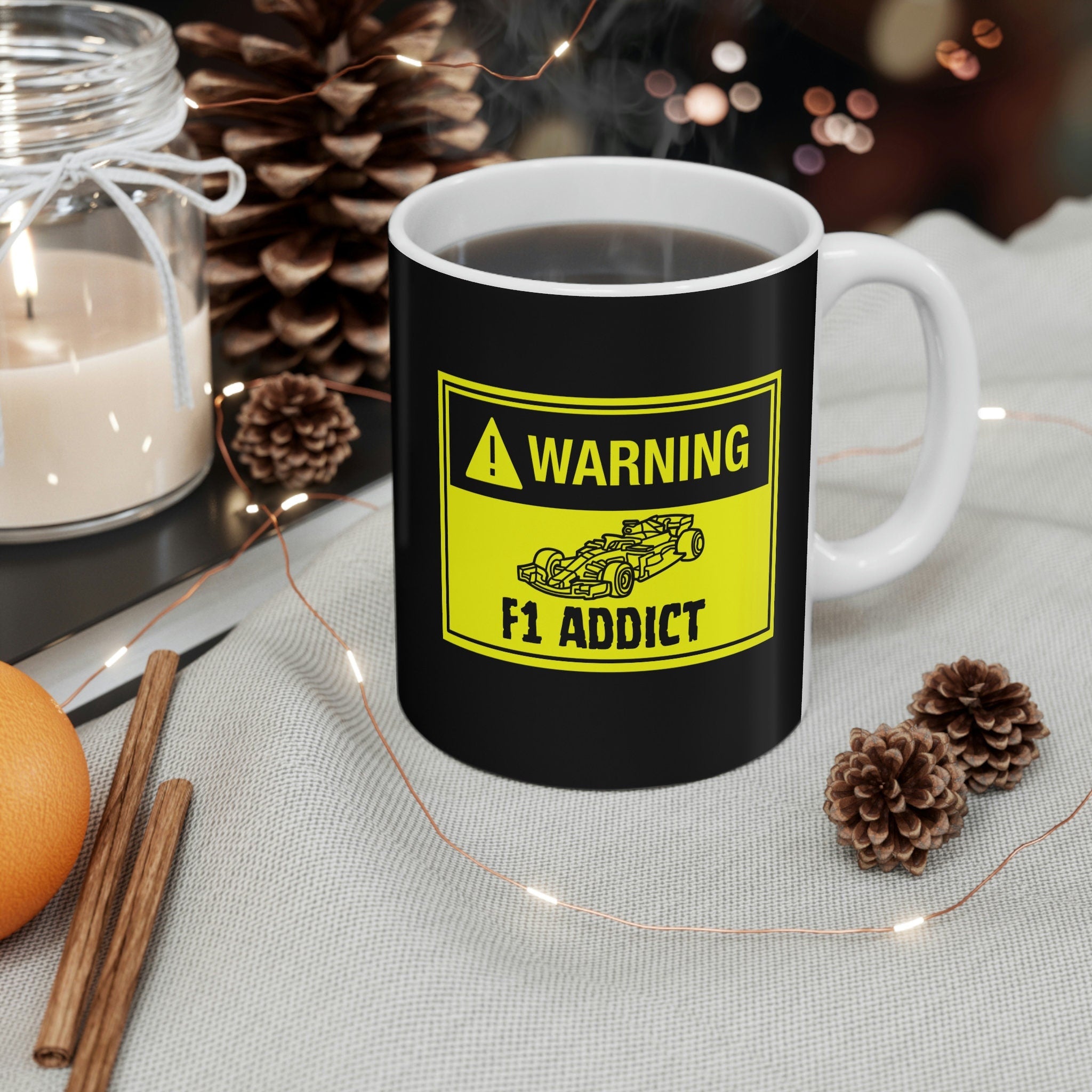 A black coffee mug with a yellow warning sign graphic. The sign says &#39;WARNING F1 ADDICT&#39; with a cartoon drawing of a Formula 1 race car. The mug is sitting on a beige surface with string lights and pine cones.