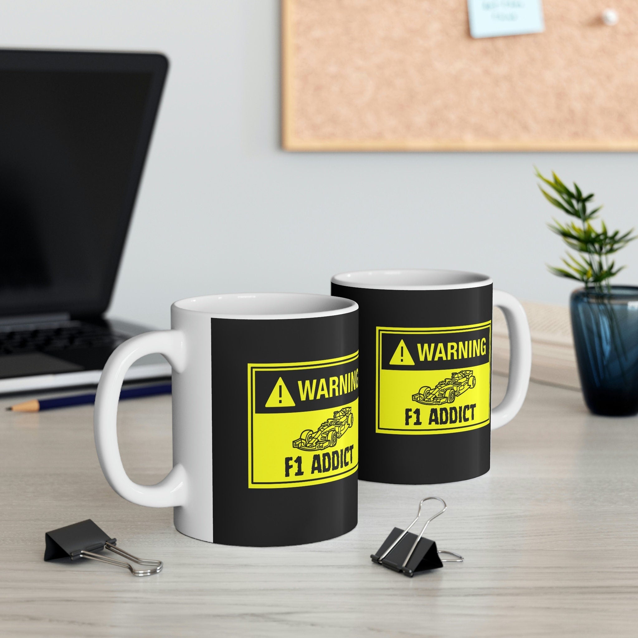 Two 11-ounce white ceramic mugs with black bodies. Each mug displays a yellow warning sign graphic with black text reading &#39;WARNING! F1 Addict.&#39;  A small black and white illustration of a Formula 1 race car is featured within the warning sign. The mugs are positioned on a light-colored wooden surface, alongside a laptop, pencils, and paper clips. The mugs are suitable for coffee, tea, or other hot beverages.