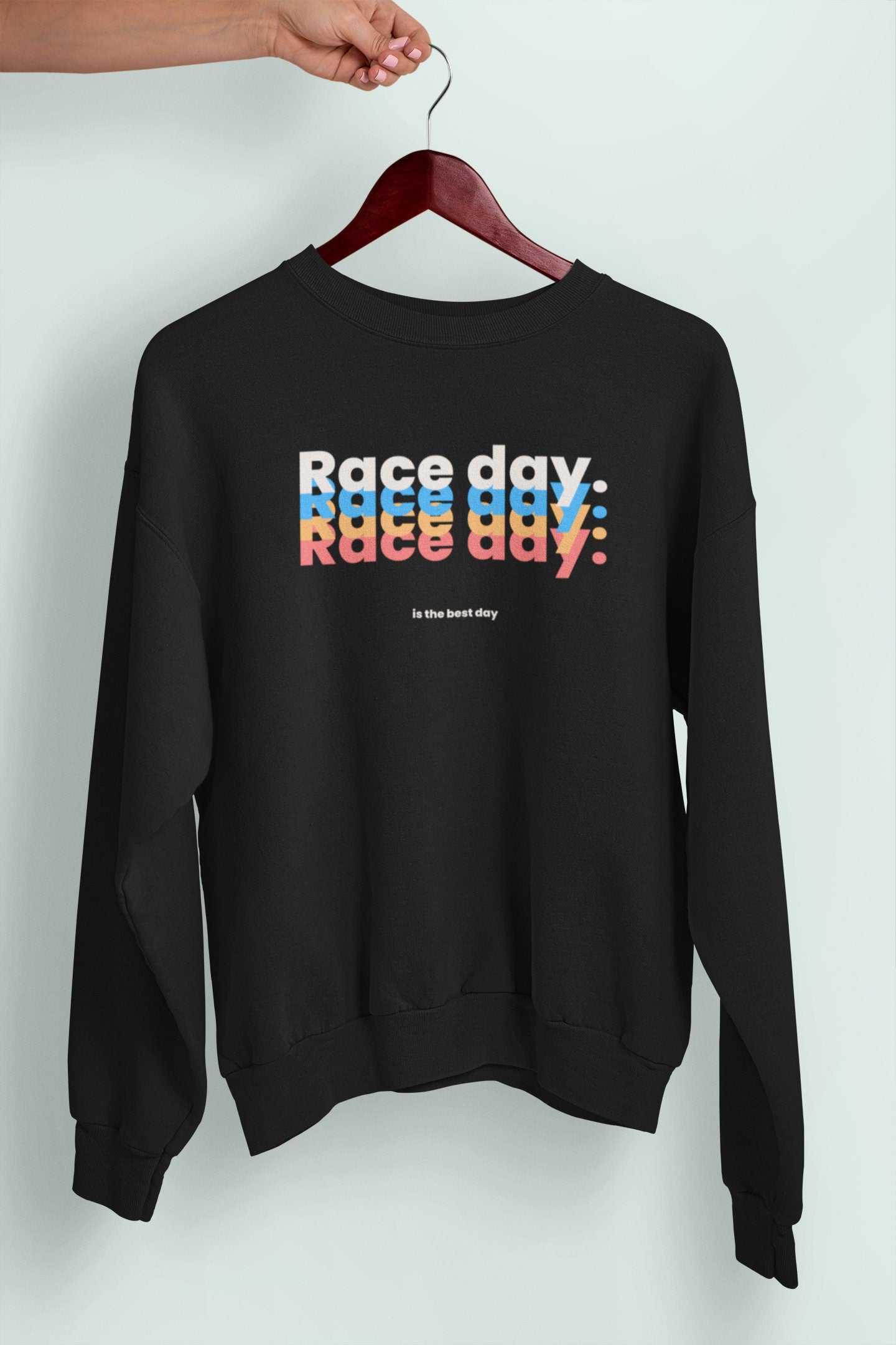 May include: Black sweatshirt with the text &#39;Race day&#39; repeated three times in stacked layers, with each layer in a different color: white, blue, and orange. The text is followed by the phrase &#39;is the best day&#39; in a smaller font size.