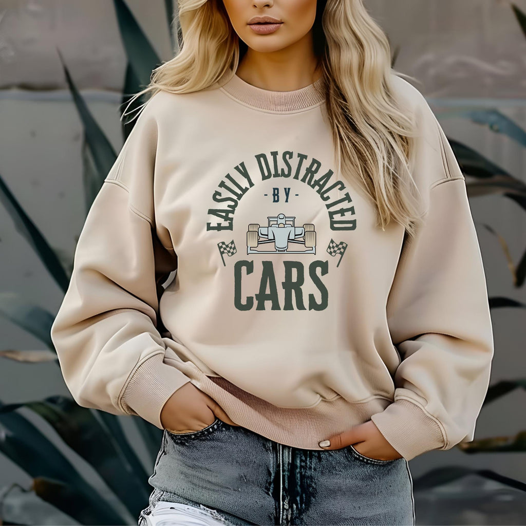 A beige sweatshirt with the text &#39;Easily Distracted - By - Cars&#39; in green lettering. The text is surrounded by a circle with a small cartoon image of a race car inside the circle.