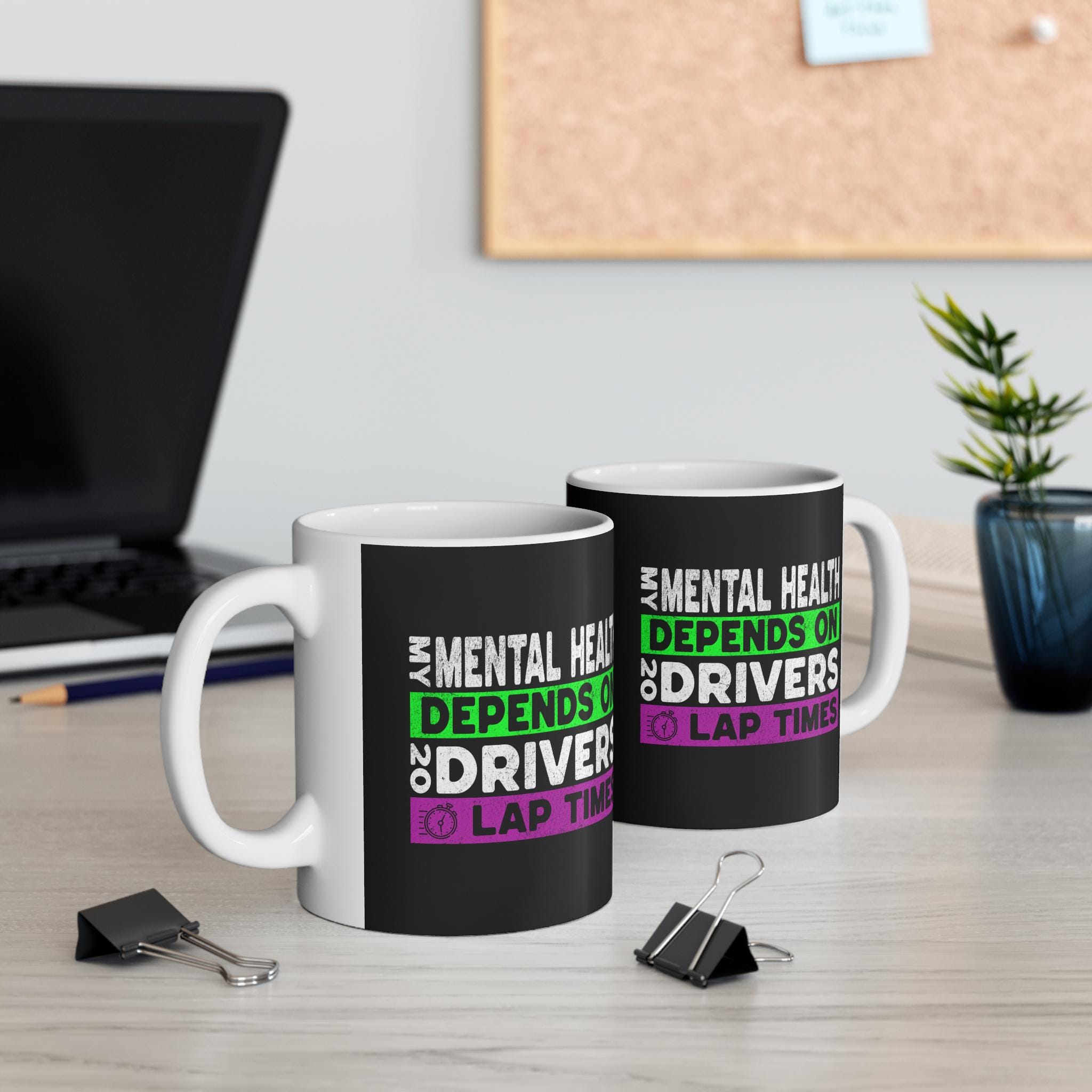 Two mugs have black backgrounds with white text that reads &#39;My Mental Health Depends On 20 Drivers Lap Times&#39; in green and purple text. The text on the left mug is slightly cut off. The mugs are sitting on a wooden surface.