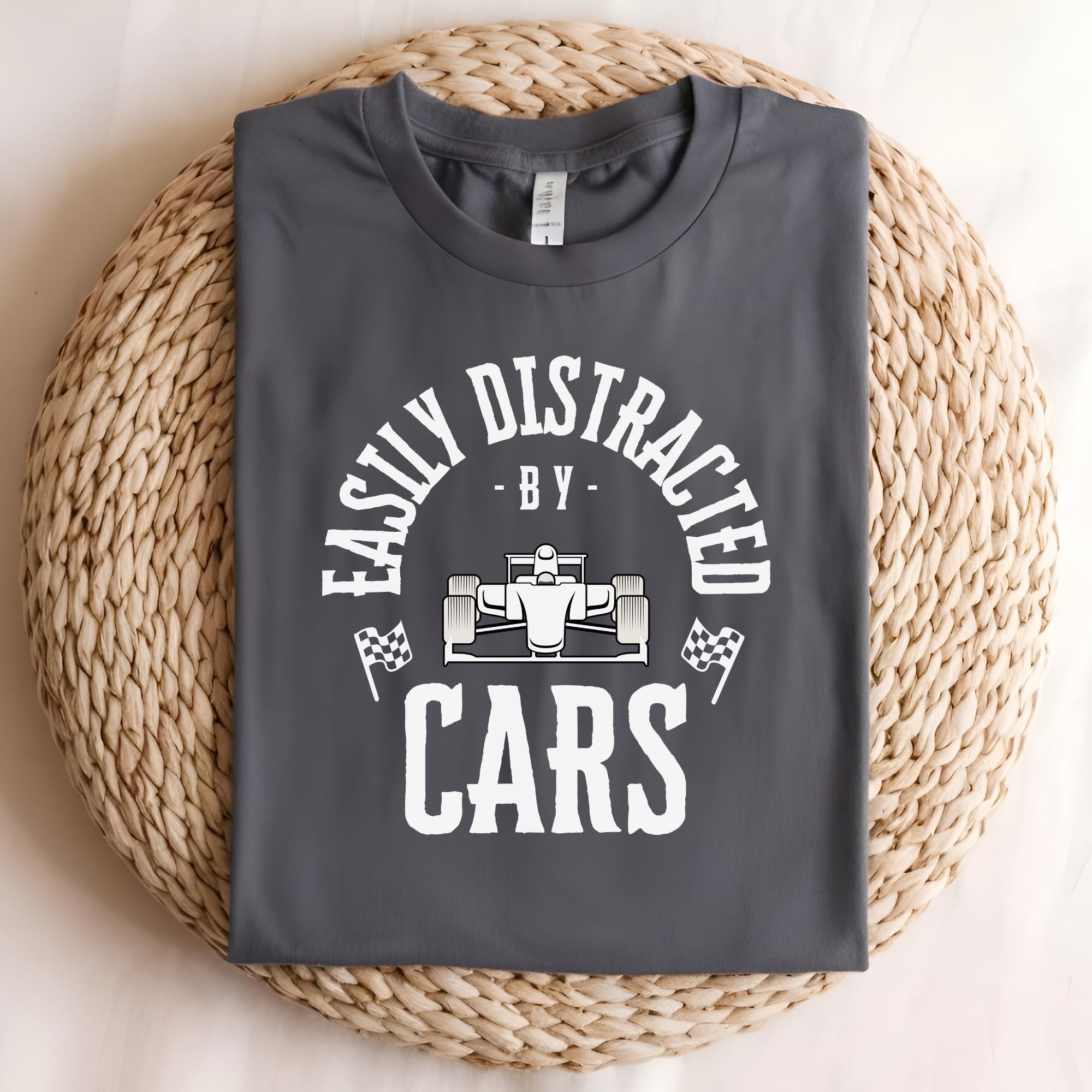 A gray t-shirt with the text &#39;Easily Distracted - By - Cars&#39; in white. A white cartoon race car is between the words &#39;By&#39; and &#39;Cars&#39;. The text is in a circle with a white checkered flag on each side of the circle.
