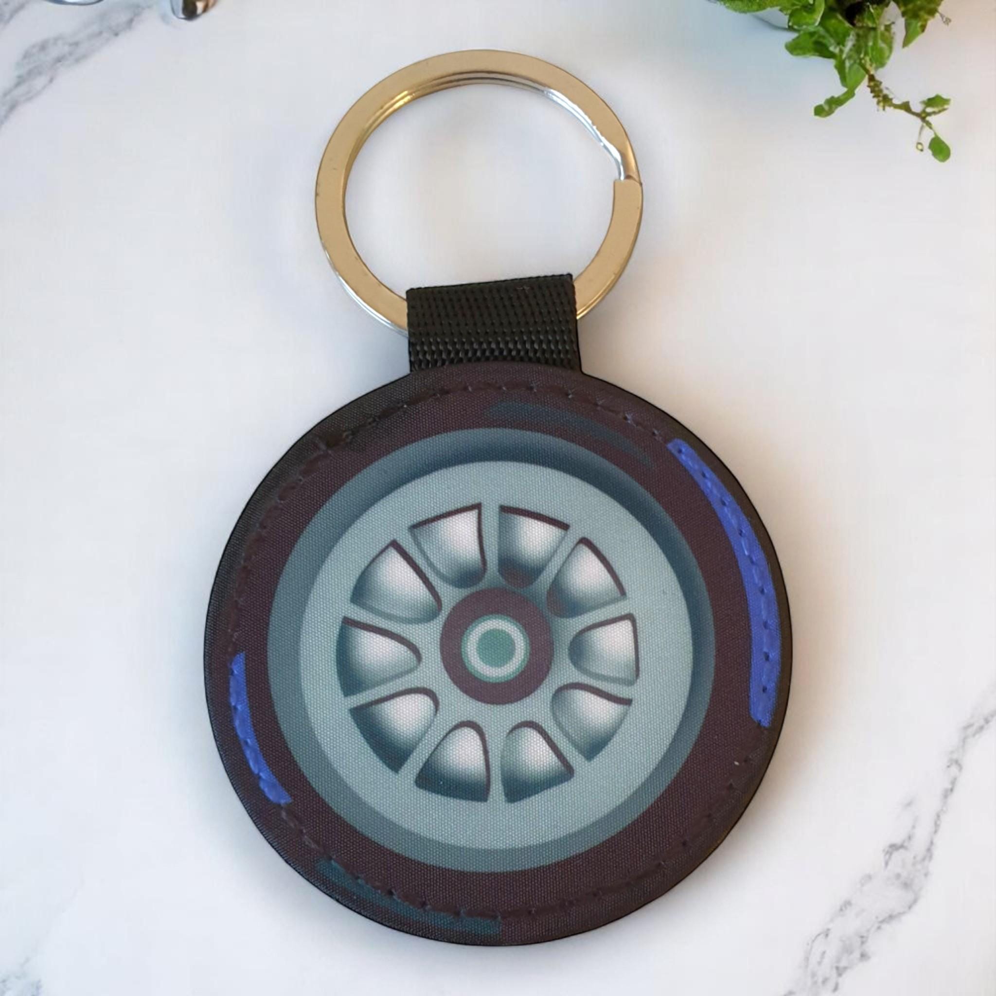 A black and gray round keychain with a blue rim. The keychain has a picture of a gray car tire with a silver rim and a black center.