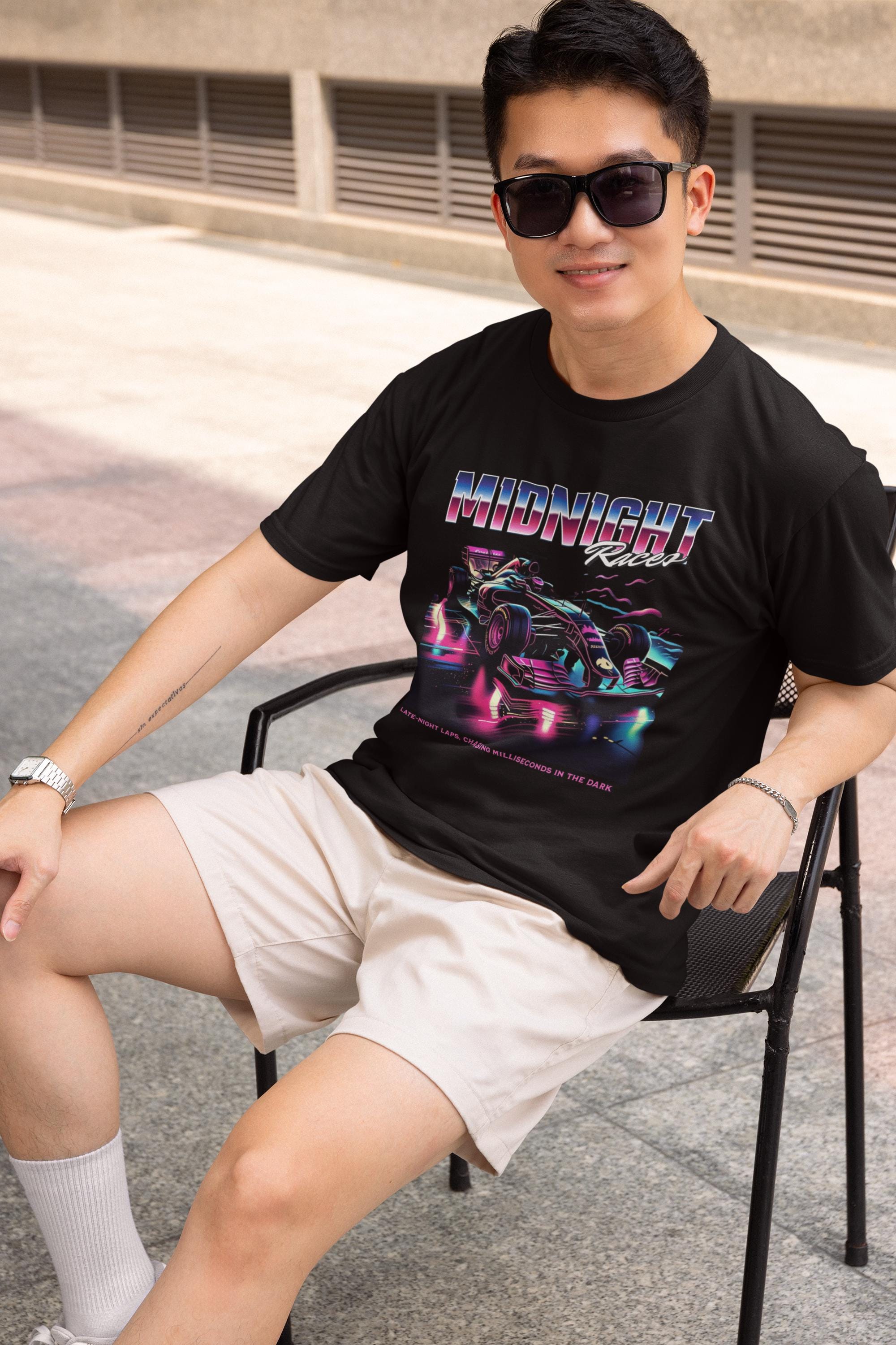 May include: A person wearing a black t-shirt with a graphic of a race car and the text &#39;MIDNIGHT Racer&#39; in a retro style. The text below the graphic reads &#39;TAKE EVERY LAP, EVERY TURN, EVERY MILLISECONDS IN THE DARK.&#39; The person is sat on a chair wearing cream shorts.