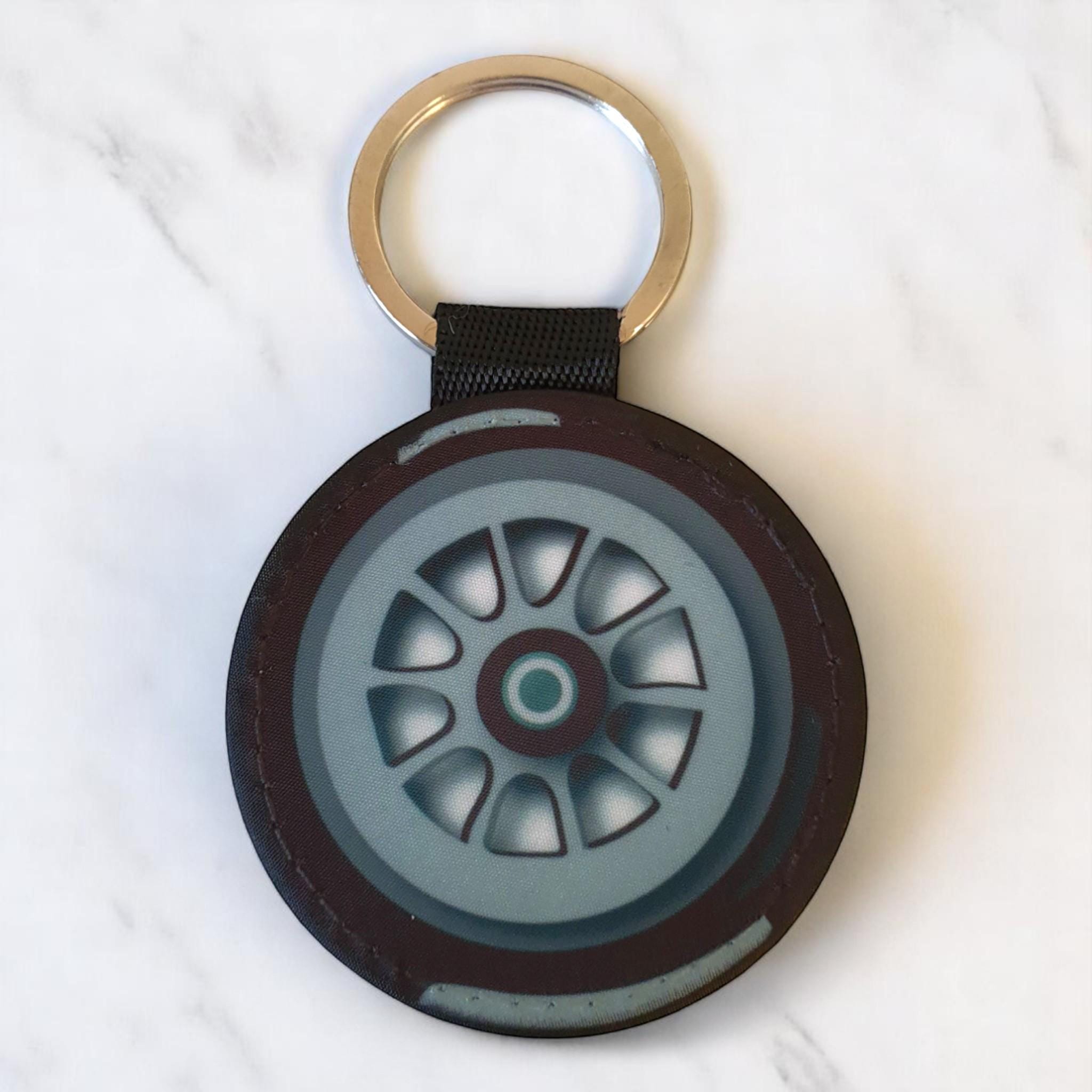 May include: A black and gray tire keychain with a silver metal ring. The tire has a detailed design of a wheel with spokes and a center cap.