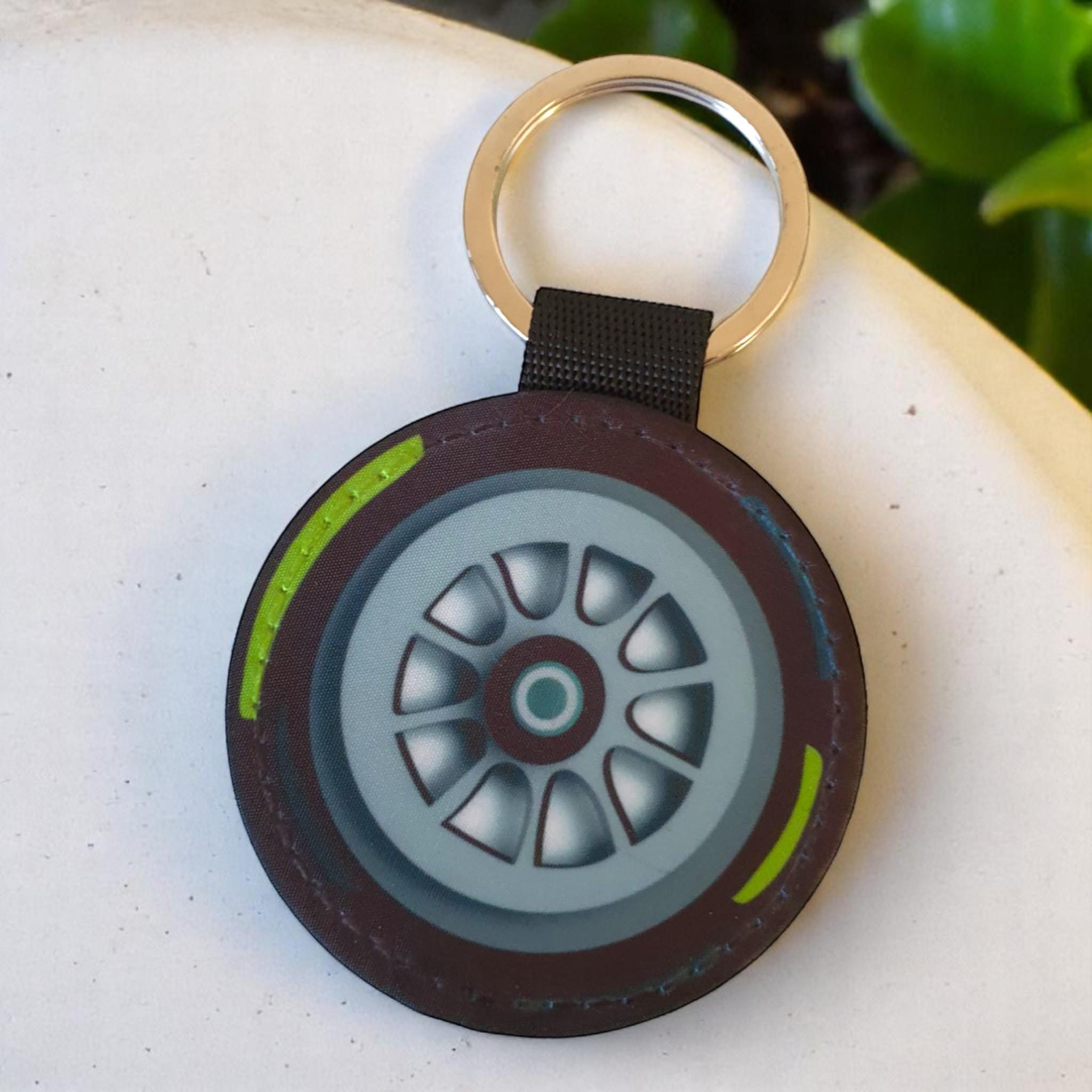 A black and brown leather keychain with a silver key ring. The keychain features a round, flat, brown leather tag with a green and white tire tread design. The tire tread design includes a silver rim with a gray tire and a yellow stripe around the edge.
