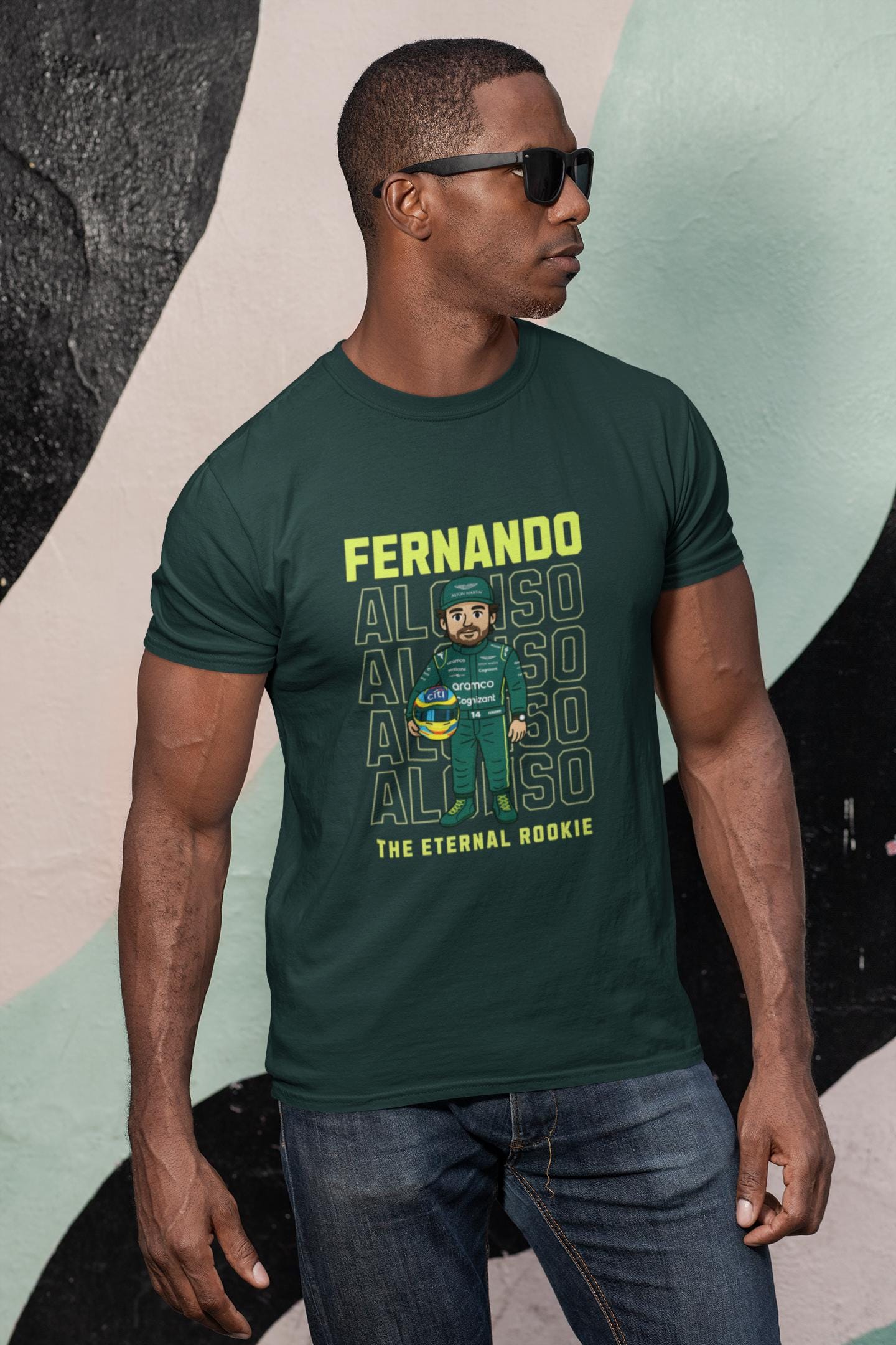 A dark green t-shirt featuring a cartoon of a Formula 1 driver in a green racing suit and helmet. The shirt has the name &#39;FERNANDO&#39; in yellow block letters above the cartoon. Below the name, the word &#39;ALONSO&#39; is repeated four times in a vertical stack. The text &#39;THE ETERNAL ROOKIE&#39; is printed below the cartoon.