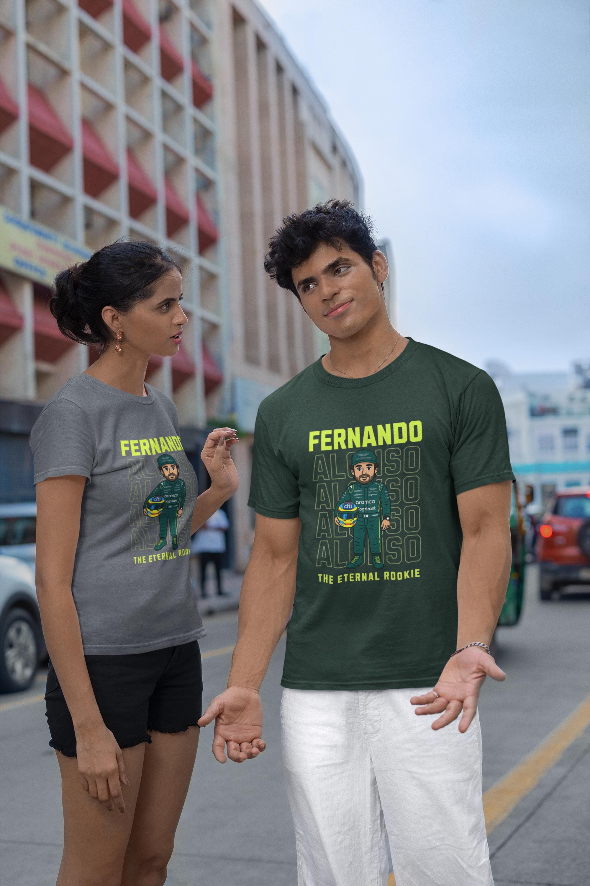 Two t-shirts, one gray and one dark green, featuring a cartoon graphic of a Formula 1 driver. The green shirt displays &#39;FERNANDO ALONSO&#39; in yellow text, with the driver&#39;s image below and &#39;THE ETERNAL ROOKIE&#39; in yellow. The gray shirt has a similar design.
