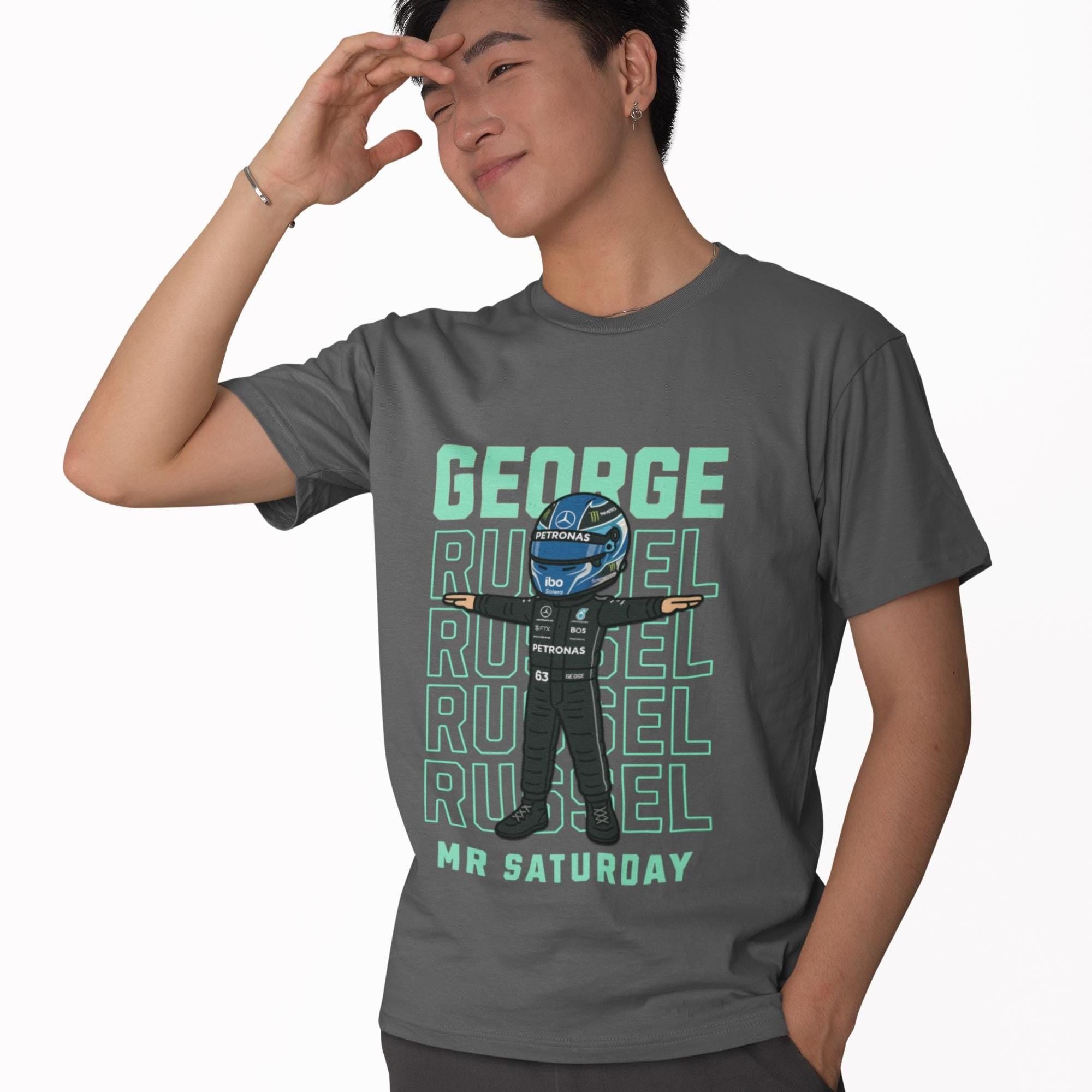 A gray t-shirt featuring a cartoon illustration of a Formula 1 driver in a racing suit and helmet. The design includes the words &#39;GEORGE RUSSEL&#39; repeated multiple times in a teal font, with &#39;MR SATURDAY&#39; below. The driver illustration is in the center, with arms outstretched.