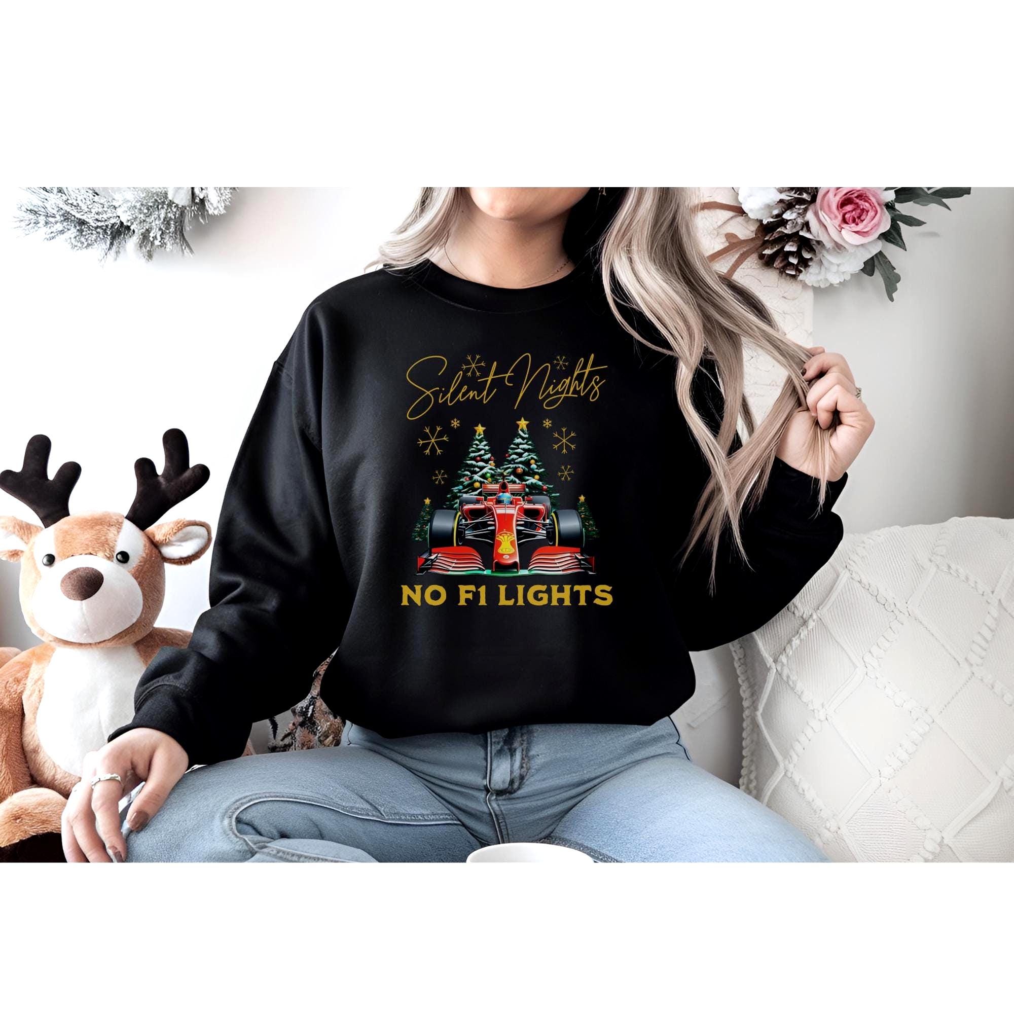 A woman wearing a black sweatshirt sits in a festive room. The sweatshirt showcases a vibrant red F1 car surrounded by decorated Christmas trees and golden snowflakes, with the words &quot;Silent Nights, No F1 Lights&quot;blending motorsport and holiday vibes.